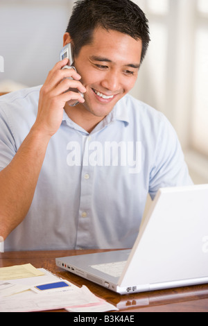 Man in dining room on cellular phone using laptop smiling Stock Photo