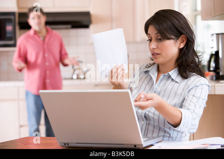 Woman in kitchen with paperwork using laptop with man in background Stock Photo