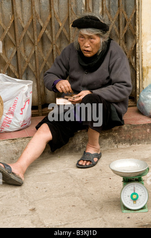 An elderly woman rolls herself a cigarette while sitting on a shop doorway in Hanoi Vietnam Stock Photo