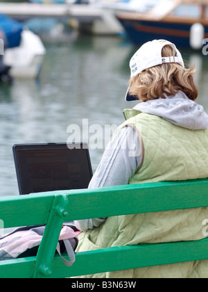 Young person sat on a bench beside Padstow harbour, typing on a laptop. Stock Photo