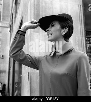 Fashions taken during London Fashion Week 1964 Wearing large peaked cap and dress with high collar with her hand raised to the peak of her hat smiling Stock Photo