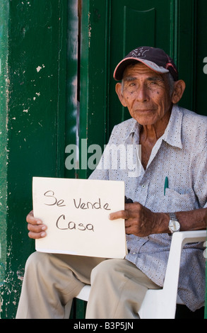 An elderly man sits in his doorway attempting to sell his house to passers by He is holding a sign that sez house for sale The e Stock Photo