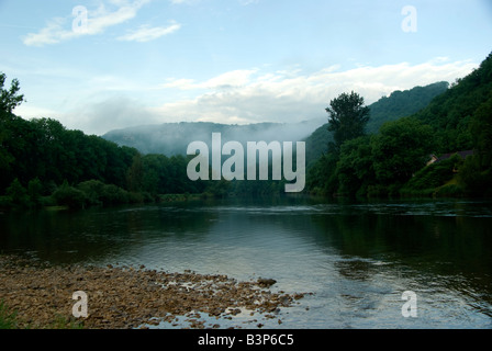 A river viewed from the stoney shallows Wooded hills rise up beyond the river and mist clings to the tree tops Stock Photo