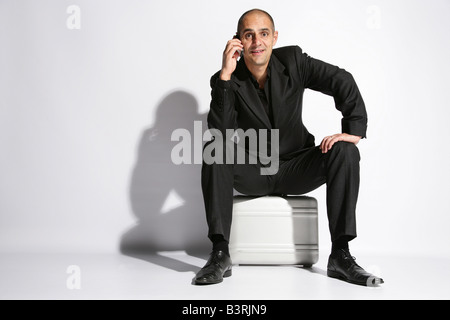 Man in a busines suit sat on a brief case holding a mobile phone Stock Photo