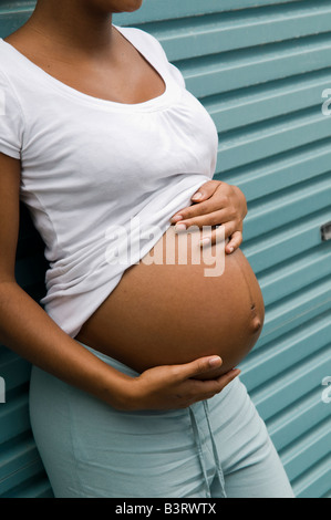 Pregnant black unmarried teenage single mother girl alone holding her big belly, leaning against blue metal garage door