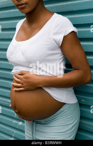 Pregnant black unmarried teenage single mother girl alone, UK, with her swollen belly exposed between jeans and white t-shirt