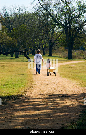 Woman and little girl pulling wagon walking side by side on pathway in public park Stock Photo