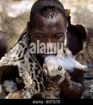 Tanzania, Northern Tanzania, Lake Eyasi. A Hadza hunter wearing a genet cat skin cape smokes cannabis from a crude stone pipe sheathed in leather.The Hadzabe are a thoUSAnd-strong community of hunter-gatherers who have lived in the Lake Eyasi basin for centuries. They are one of only four or five societies in the world that still earn a living primarily from wild resources. . Stock Photo