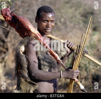 Tanzania, Northern Tanzania, Lake Eyasi. A Hadza hunter wearing a baboon skin returns to camp with a haunch of impala over his shoulder. He killed the antelope with a metal-tipped arrow that had been dipped in a fast-acting vegetable poison extracted from the desert rose (Adenium obesum).The Hadzabe are a thoUSAnd-strong community of hunter-gatherers who have lived in the Lake Eyasi basin for centuries. They are one of only four or five societies in the world that still earn a living primarily from wild resources.. . Stock Photo