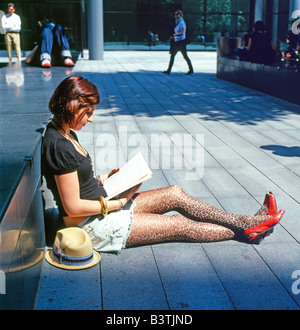 A young woman wearing red shoes sitting on the street sidewalk pavement  reading book Bishops Square Spitalfields East London England UK  KATHY DEWITT Stock Photo