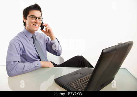Smiling Asian businessman sitting at desk talking on cellphone Stock Photo