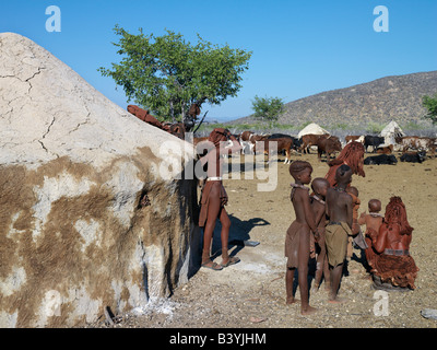 Namibia, Kaokoland. A Himba family looks at their family's cattle outside their house. The house is plastered with a mixture of Stock Photo