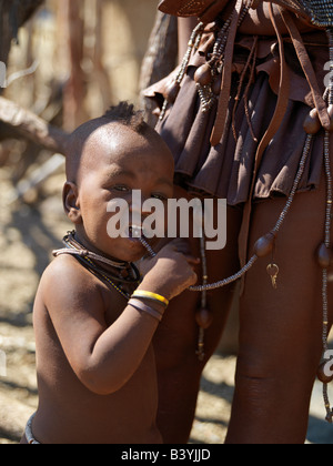 Namibia, Kaokoland. A child sucks a bead hanging from his mother's belt. A woman's body is smeared with a mixture of red ochre, Stock Photo