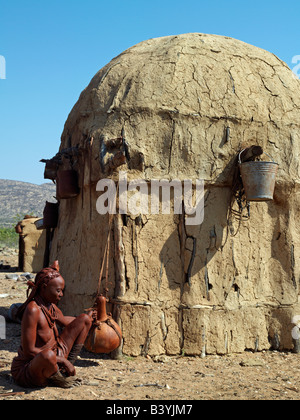 Namibia, Kaokoland. An old Himba woman gently shakes a large milk gourd to make butter outside her home. Her body gleams from a Stock Photo