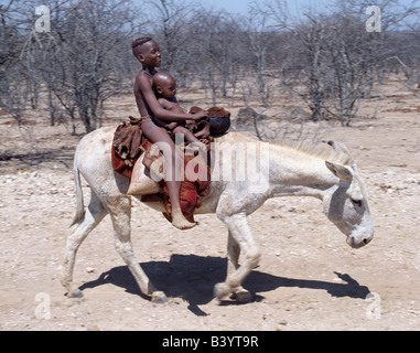 Namibia, Kaokoland, Opuwo. Two Himba children ride home on a white donkey. Their bodies gleam from a mixture of red ochre, butterfat and herbs. The rounded hairstyle of the elder child, a boy, is typical of Himba youth.The Himba are Herero-speaking Bantu nomads who live in the harsh, dry but starkly beautiful landscape of remote northwest Namibia. Stock Photo