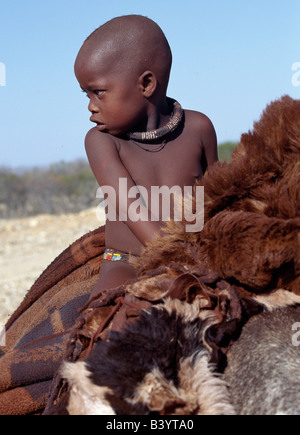 Namibia, Kaokoland, Opuwo. A young Himba boy rides home comfortably on a donkey. Sheepskins and blankets make up his saddle. His body is smeared with a mixture of red ochre, butterfat and herbs. He already wears a traditional white-beaded necklace, called ombwari, which will become larger and heavier as he grows older.The Himba are Herero-speaking Bantu nomads who live in the harsh, dry but starkly beautiful landscape of remote northwest Namibia. Stock Photo