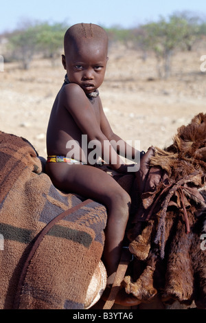 Namibia, Kaokoland, Opuwo. A young Himba boy rides home comfortably on a donkey. Sheepskins and blankets make up his saddle. His body is smeared with a mixture of red ochre, butterfat and herbs. He already wears a traditional white-beaded necklace, called ombwari, which will become larger and heavier as he grows older.The Himba are Herero-speaking Bantu nomads who live in the harsh, dry but starkly beautiful landscape of remote northwest Namibia. Stock Photo