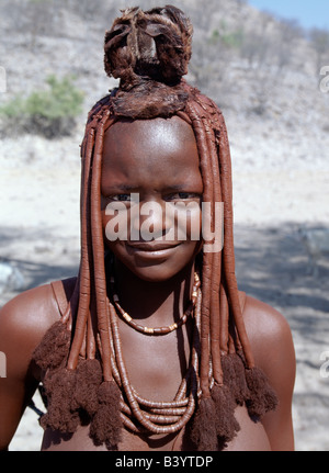 Namibia, Kaokoland, Epupa. A young Himba girl in traditional attire. Her body gleams from a mixture of red ochre, butterfat and herbs. Her hair is styled in the traditional Himba way and is crowned with a headdress made of lambskin, called erembe. Her necklace includes ostrich-shell beads. The Himba are Herero-speaking Bantu nomads who live in the harsh, dry but starkly beautiful landscape of remote northwest Namibia. Stock Photo