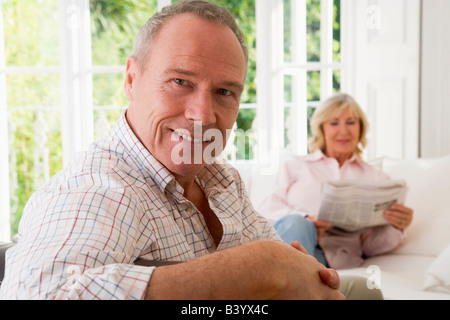 Man in living room smiling with woman in background reading newspaper Stock Photo