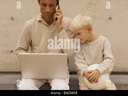 Man using a laptop while phoning with a mobile phone, boy with ball sitting next to him Stock Photo