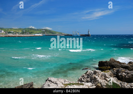 Sea aquamarine waves dash against the wind, azure sky, remote green hills and pier with lighthouse in the background Stock Photo
