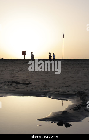 A family of three silhouetted by the setting sun walk past a red flag and lifebouy and a seawater pool at dusk on the beach Stock Photo