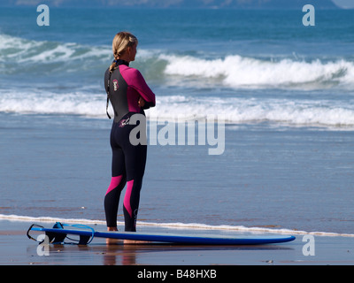 Blonde female surfer standing at the edge of the water looking at the waves. Watergate Bay, Newquay, Cornwall Stock Photo