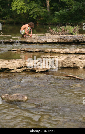 A young boy kneels on a rock in the middle of the Little Harpeth River in Nashville, as he makes a raft of sticks and leaves. Stock Photo