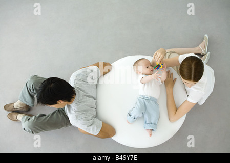 Mother playing with infant lying on ottoman, father sitting with back turned to them, overhead view Stock Photo