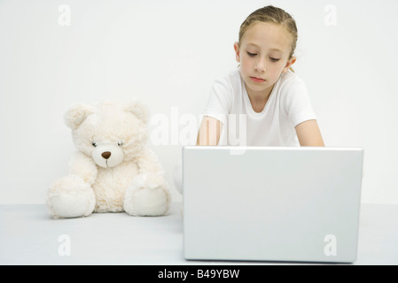 Girl sitting beside teddy bear, using laptop computer Stock Photo