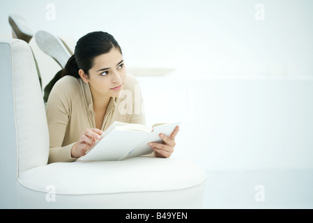 Young woman lying on stomach on sofa, holding book, looking away Stock Photo