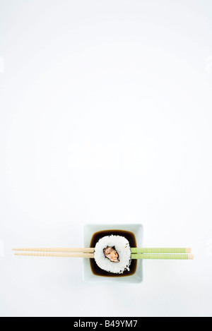 Single piece of maki sushi on chopsticks over dish of soy sauce, overhead view Stock Photo