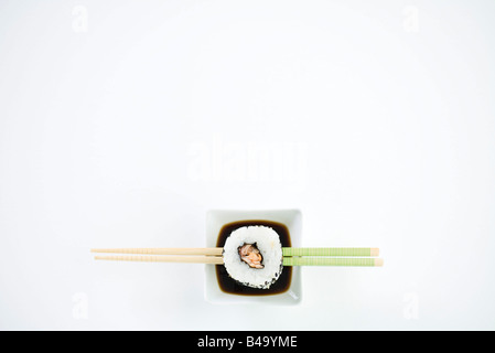 Single piece of maki sushi resting on chopsticks over dish of soy sauce, overhead view Stock Photo