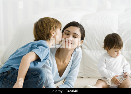 Little boy kissing mother's cheek, toddler girl sitting nearby Stock Photo