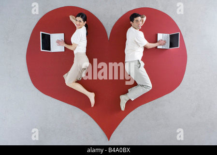 Man and woman lying on large heart graphic, both using laptop computers Stock Photo