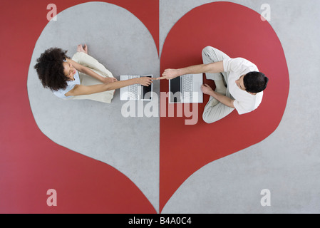 Couple sitting face to face with laptop computers on heart shape, touching fingers, overhead view Stock Photo