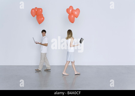 Man and woman walking past each other, both carrying laptop computers and heart balloons Stock Photo