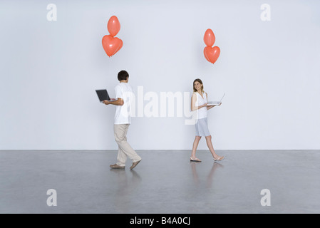 Man and woman carrying laptop computers and heart balloons, smiling over their shoulders at each other Stock Photo