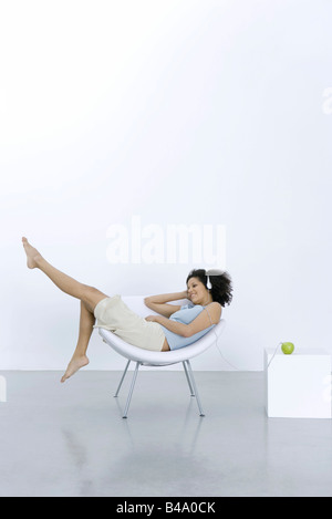 Young woman lounging in chair, listening to headphones connected to apple Stock Photo