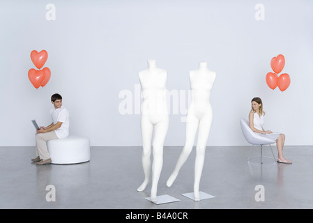 Man and woman sitting with laptops and heart balloons, looking over shoulders at each other Stock Photo