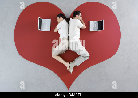 Man and woman lying back to back on large heart, both looking at laptop computers Stock Photo