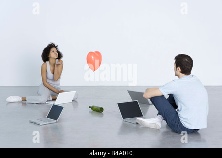 Man and woman seated with laptops, woman blowing man a kiss, bottle and heart balloon between them Stock Photo