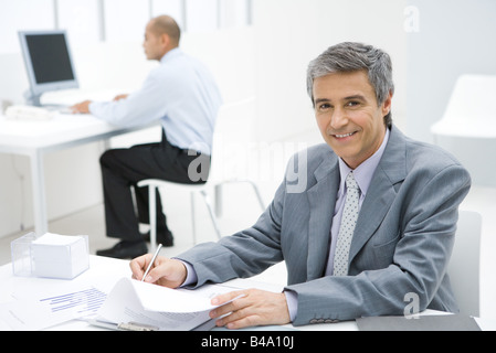 Businessman working at desk, smiling at camera, colleague in background Stock Photo