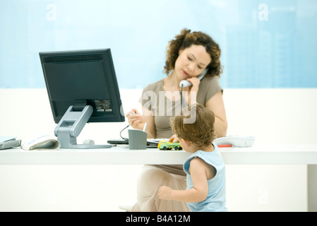 Woman using phone in office, toddler son playing with toy truck on desk Stock Photo
