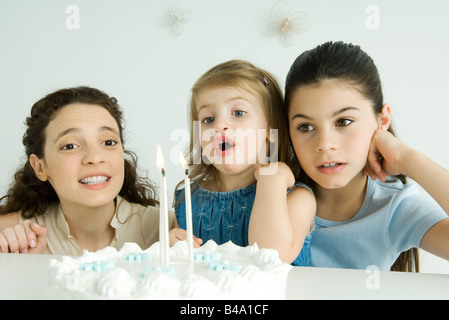 Little girl blowing out candles on birthday cake, mother and older sister watching Stock Photo