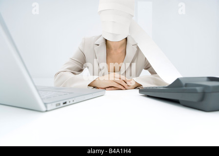 Professional woman sitting at desk, paper from adding machine wrapped around face Stock Photo