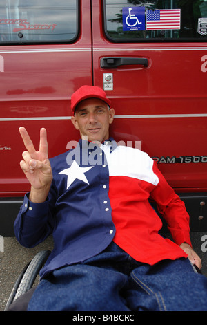 Man in a Wheelchair Gesturing Peace Sign He is Wearing a Red White and Blue Shirt with a Star on it and is Leaning Against a Van Stock Photo