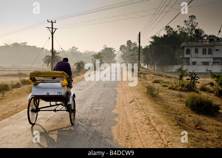 Bicycle rickshaw driver pedaling down long road in rural area. West Bengal, India. Stock Photo