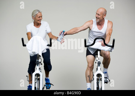 A senior woman and a mature man riding stationary bikes and passing a water bottle Stock Photo