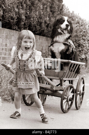 A young girl laughs out loud as she tried to pull a cart with a large dog sitting in it Stock Photo